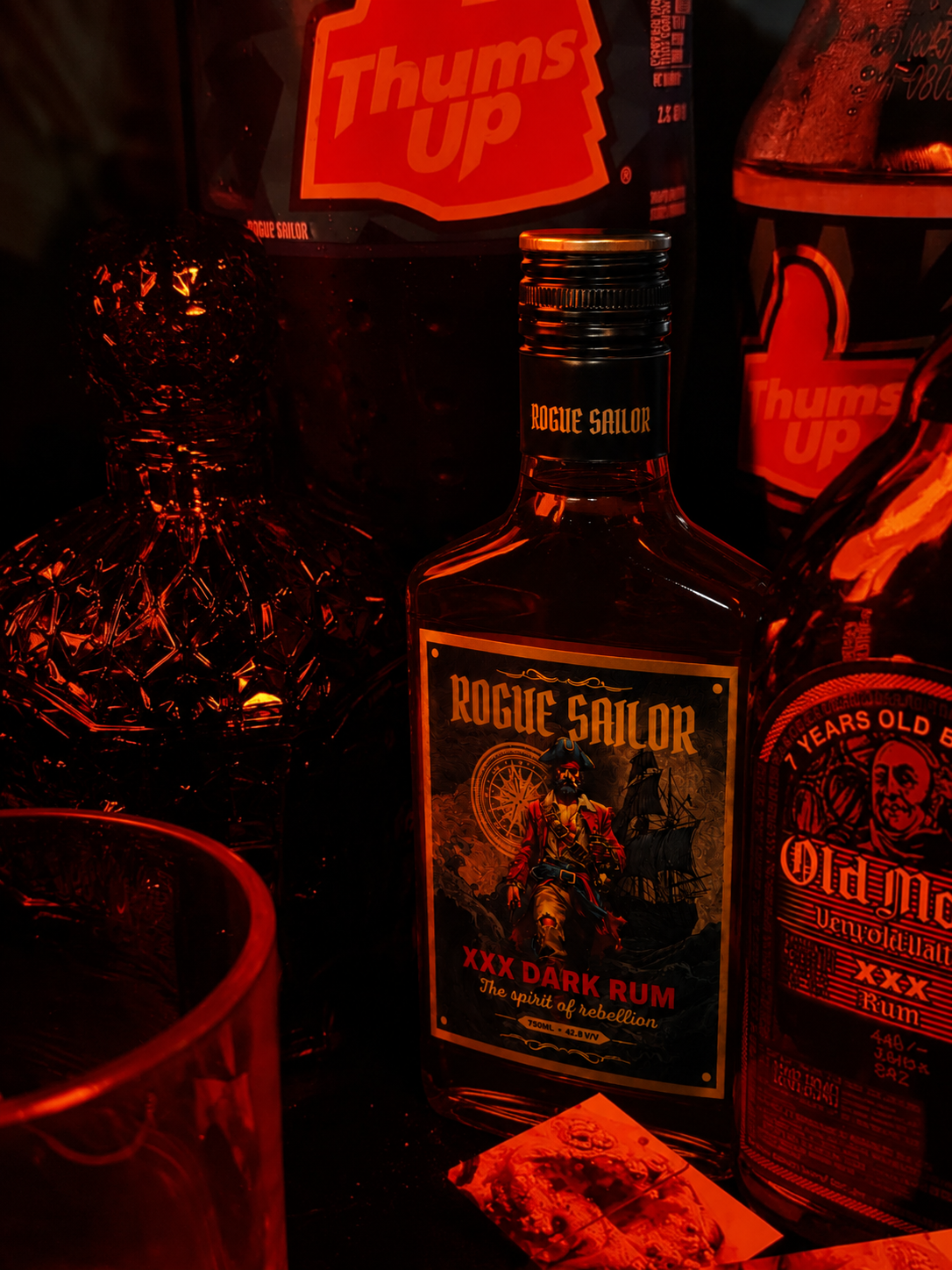 Rogue Sailor bottle in red bar light beside Thums Up and other rum bottles